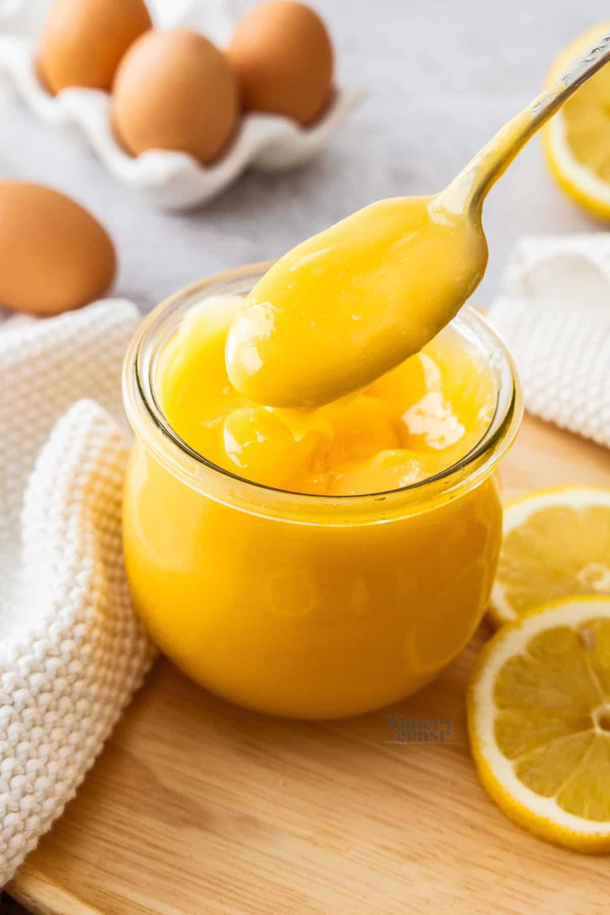 A spoon lifting lemon curd out of a glass jar.