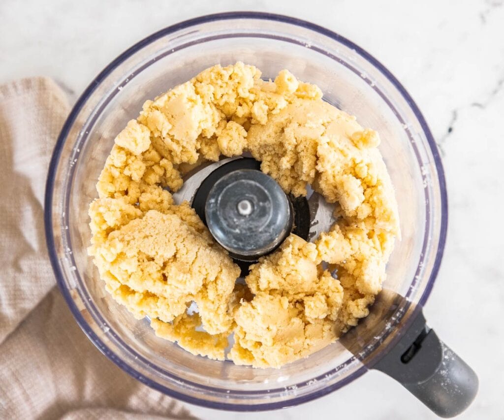 Pâte sablée dough in clumps in a food processor bowl.
