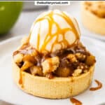An apple crumble tartlet on a dessert plate, topped with ice cream and caramel sauce.