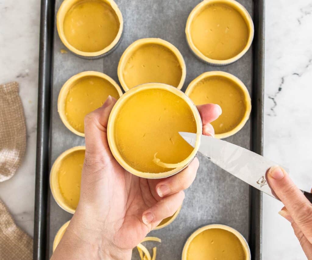 Hands using a small knife to trim away excess pastry on the tart shells.