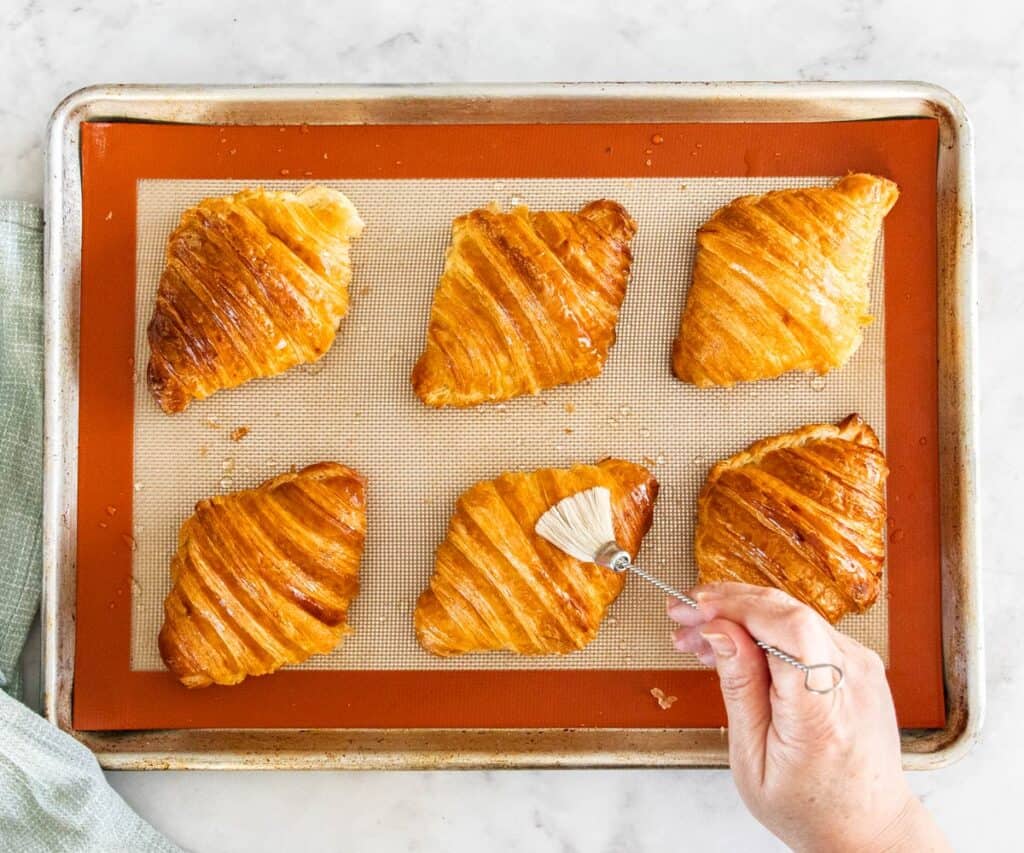 Syrup being brushed over croissants on a baking tray.