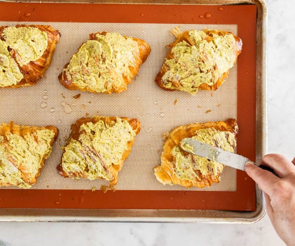 Pistachio frangipane being spread over the top of croissants on a baking tray.