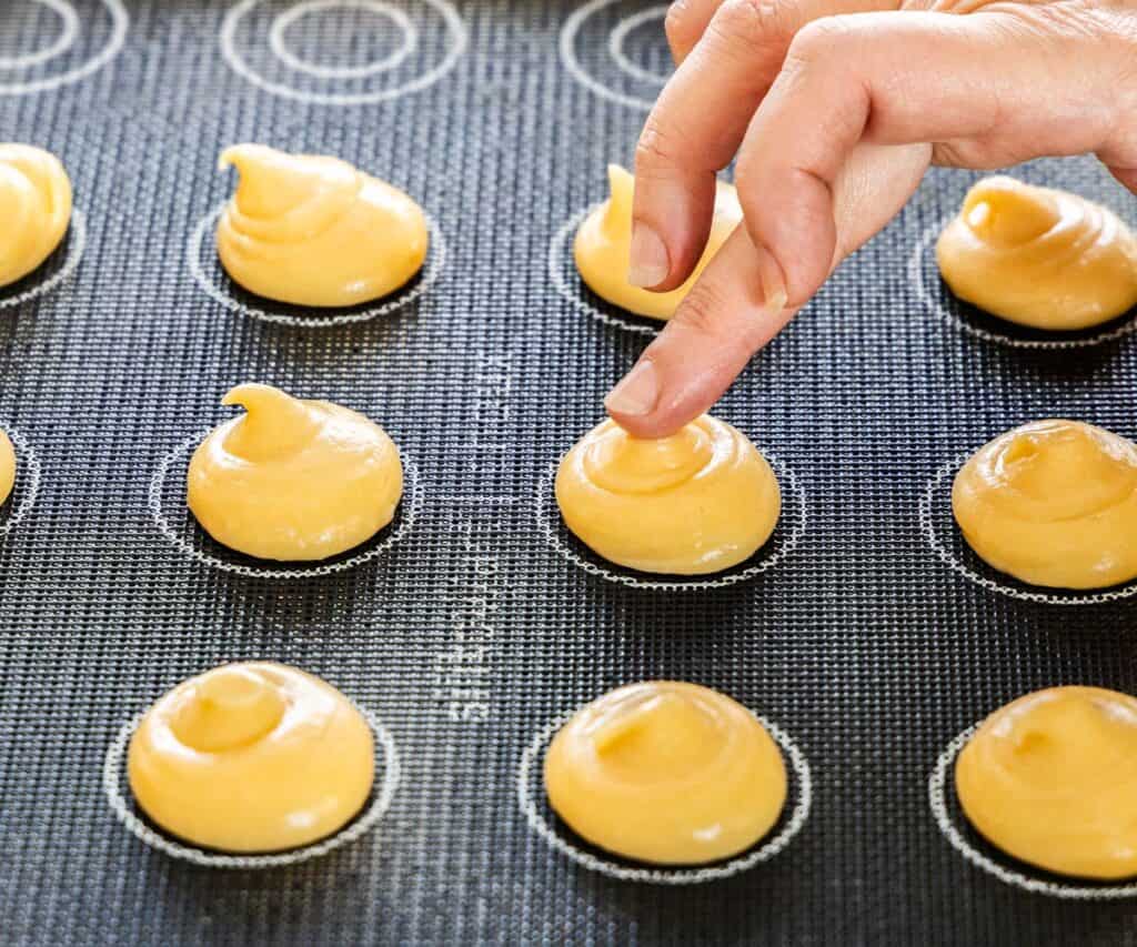 A finger pressing down the tips of the raw choux buns.