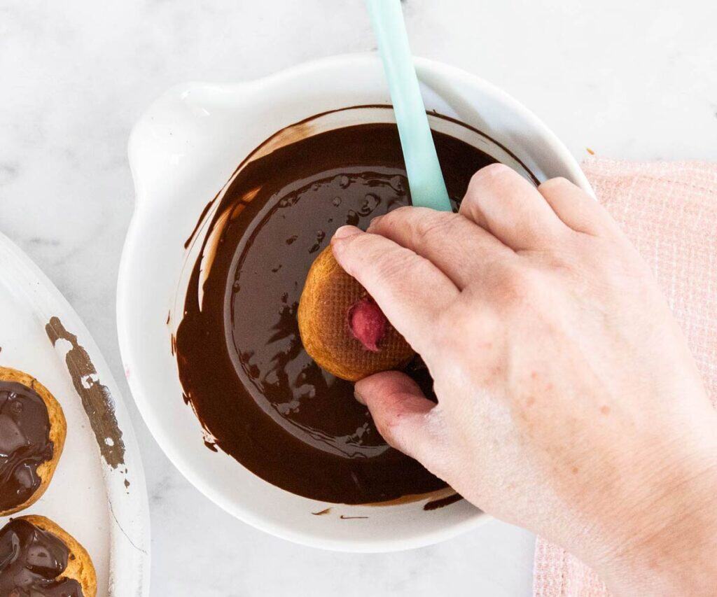 A hand dunking a profiterole in melted chocolate.