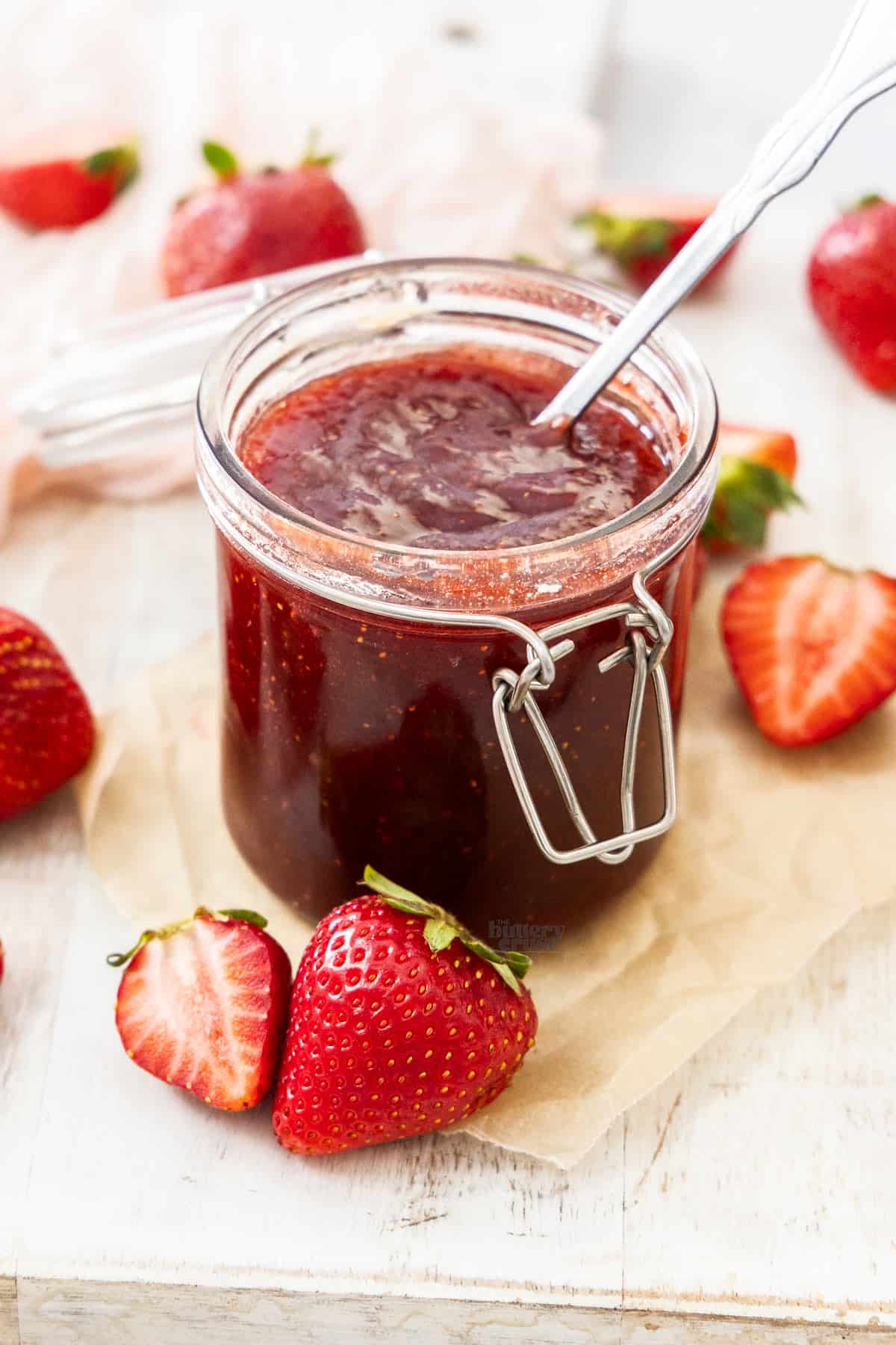 A jar of strawberry jam with a spoon sticking out, surrounded by fresh strawberries.