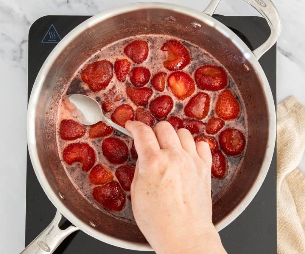 A hand using a spoon to scoop froth off the top of strawberries in a pan.
