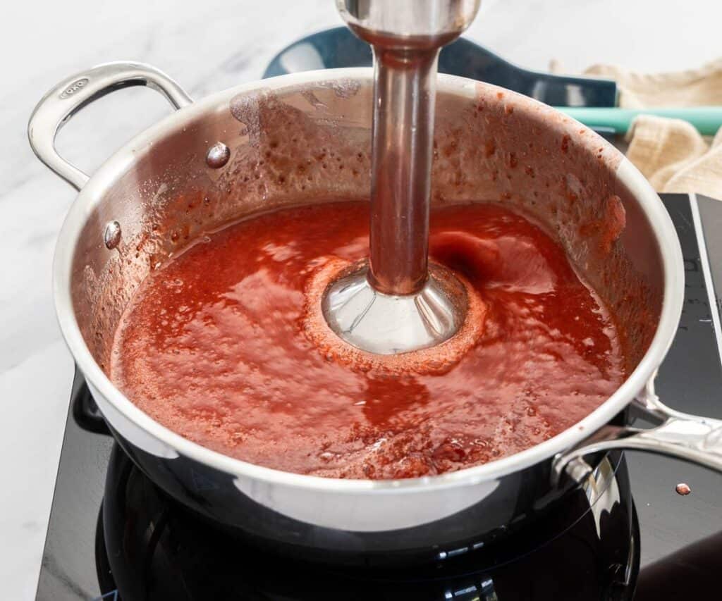 An immersion blender blending strawberries in a saucepan.