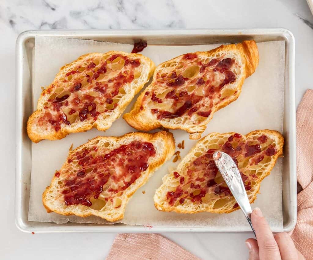 Jam being spread onto the insides of sliced croissants.