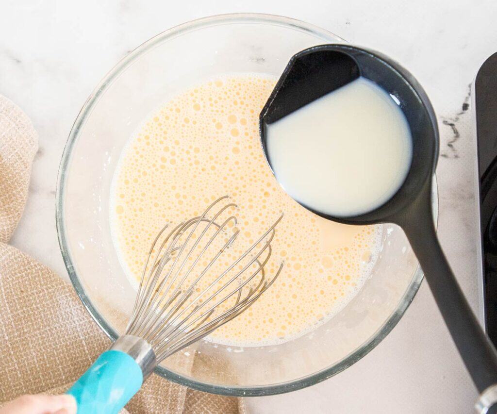 Hot milk being ladled into an egg mixture in a mixing bowl.