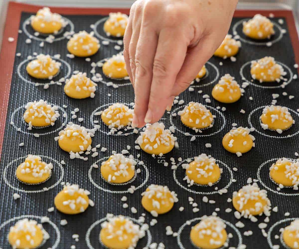 A hand scattering pearl sugar over small mounds of choux dough.