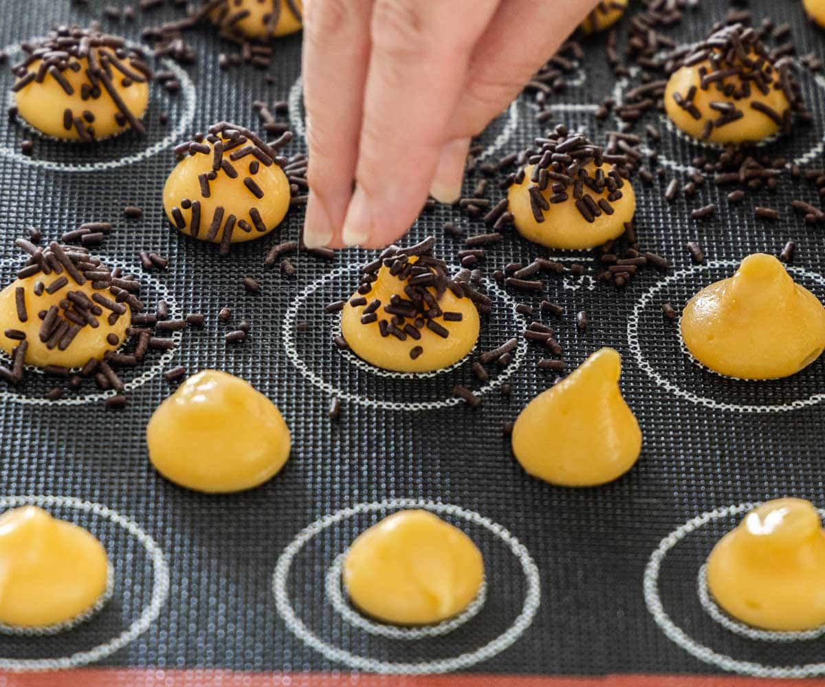 A hand sprinkling chocolate sprinkles over small mounds of choux dough.
