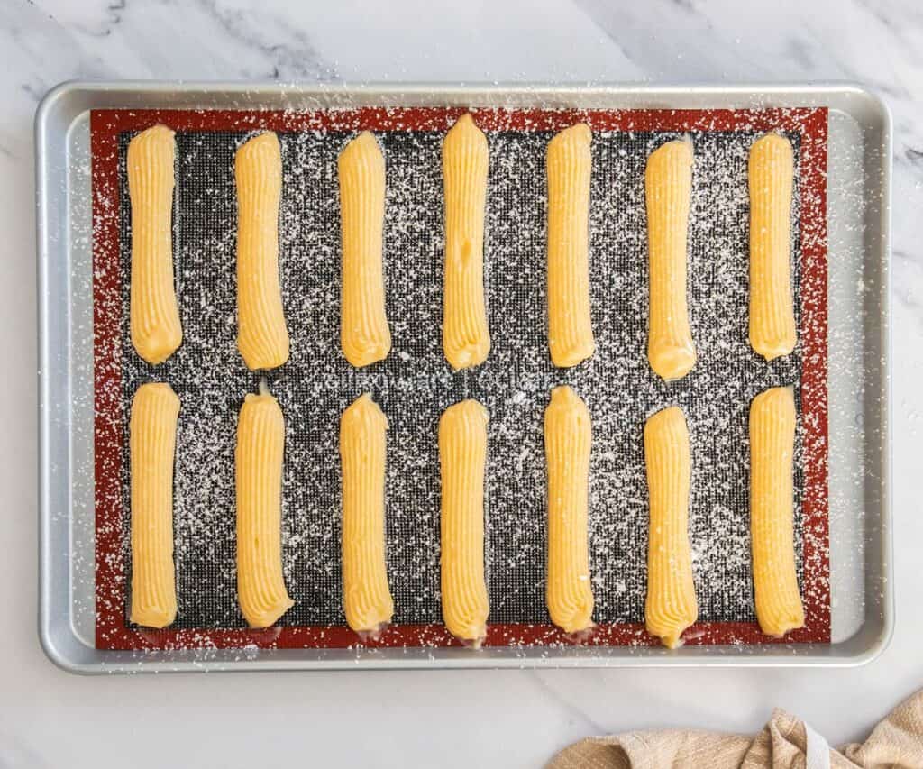 Piped eclairs, dusted with icing sugar, and ready to bake.