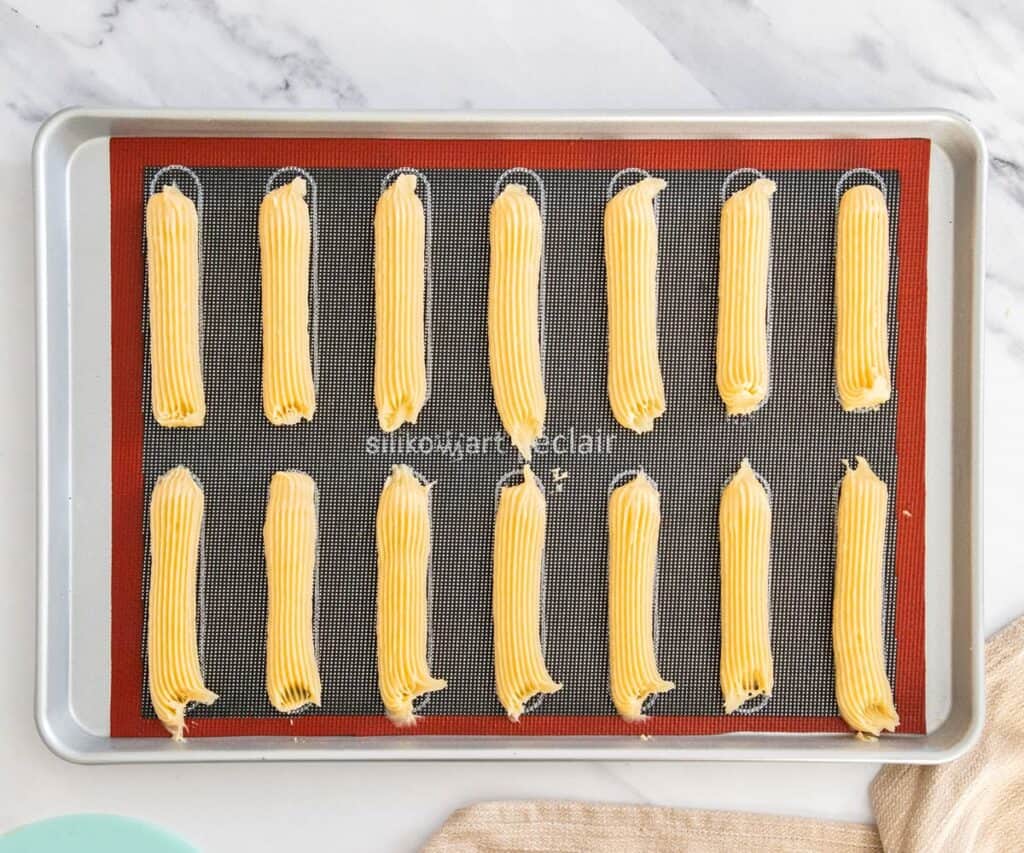 A baking sheet with a dark mat and piped eclairs on top.