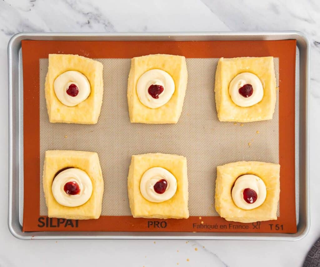 Raw danish pastry squares topped with cheesecake filling and jam.