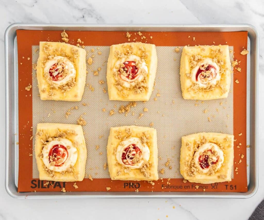 6 raw strawberry danishes on a baking tray filled with cheesecake, jam and crumble.