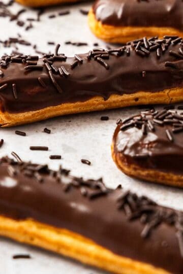 Closeup of 3 chocolate elcairs topped with, and surrounded by, chocolate sprinkles.