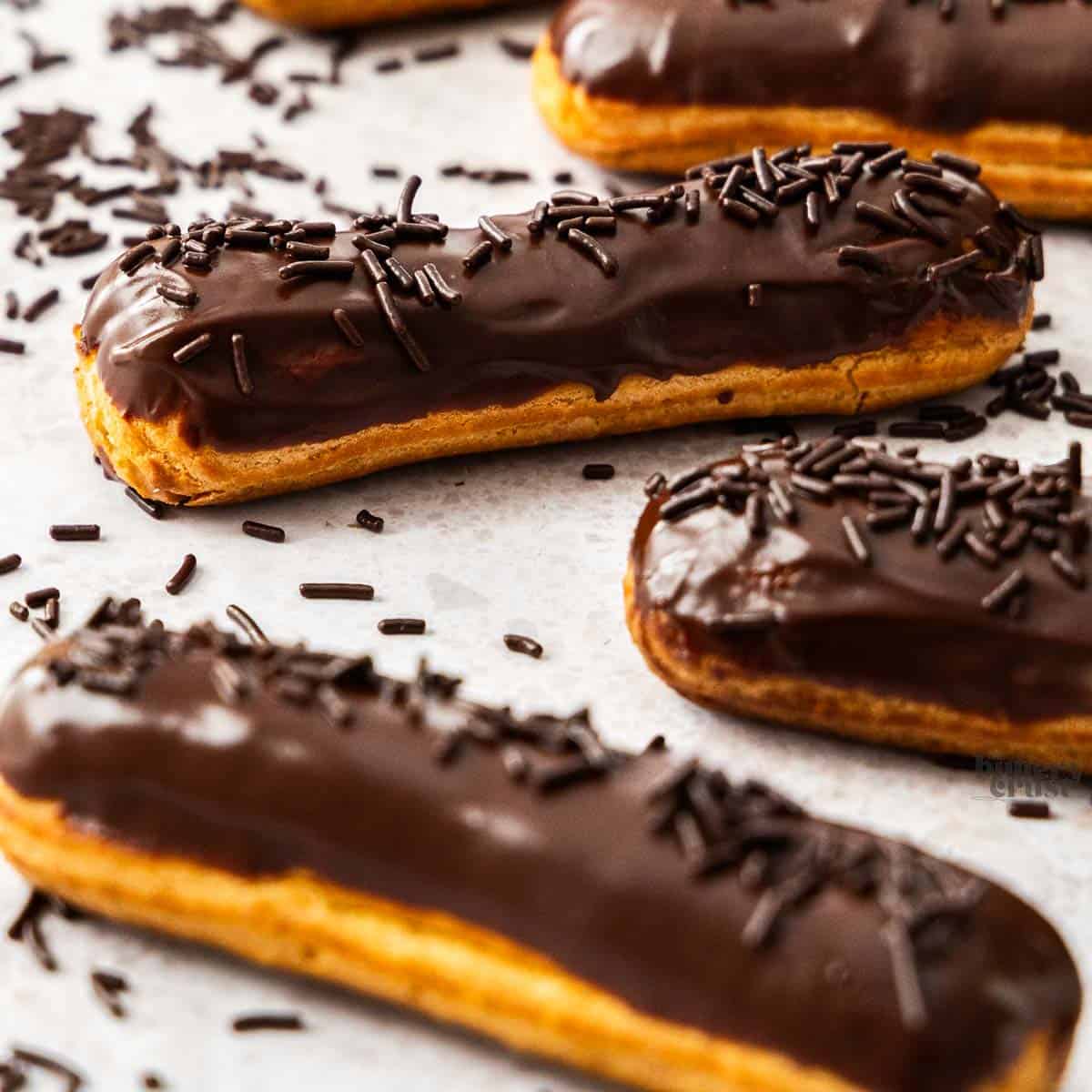 Closeup of 3 chocolate elcairs topped with, and surrounded by, chocolate sprinkles.