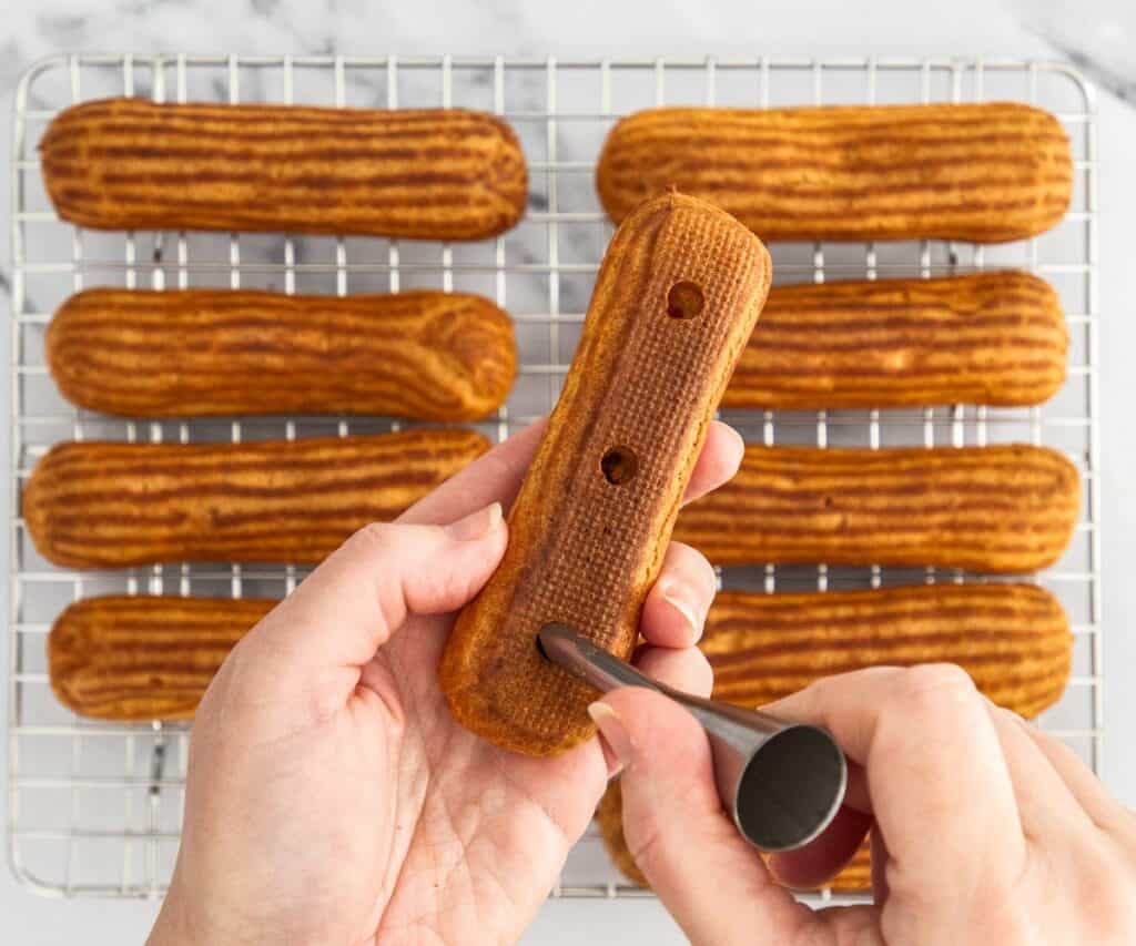 A hand holding an eclair with a piping tip in the other poking holes in the base of an eclair.