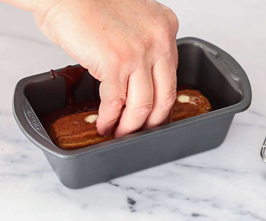 A chocolate eclair being dipped into chocolate glaze in a mini loaf pan.