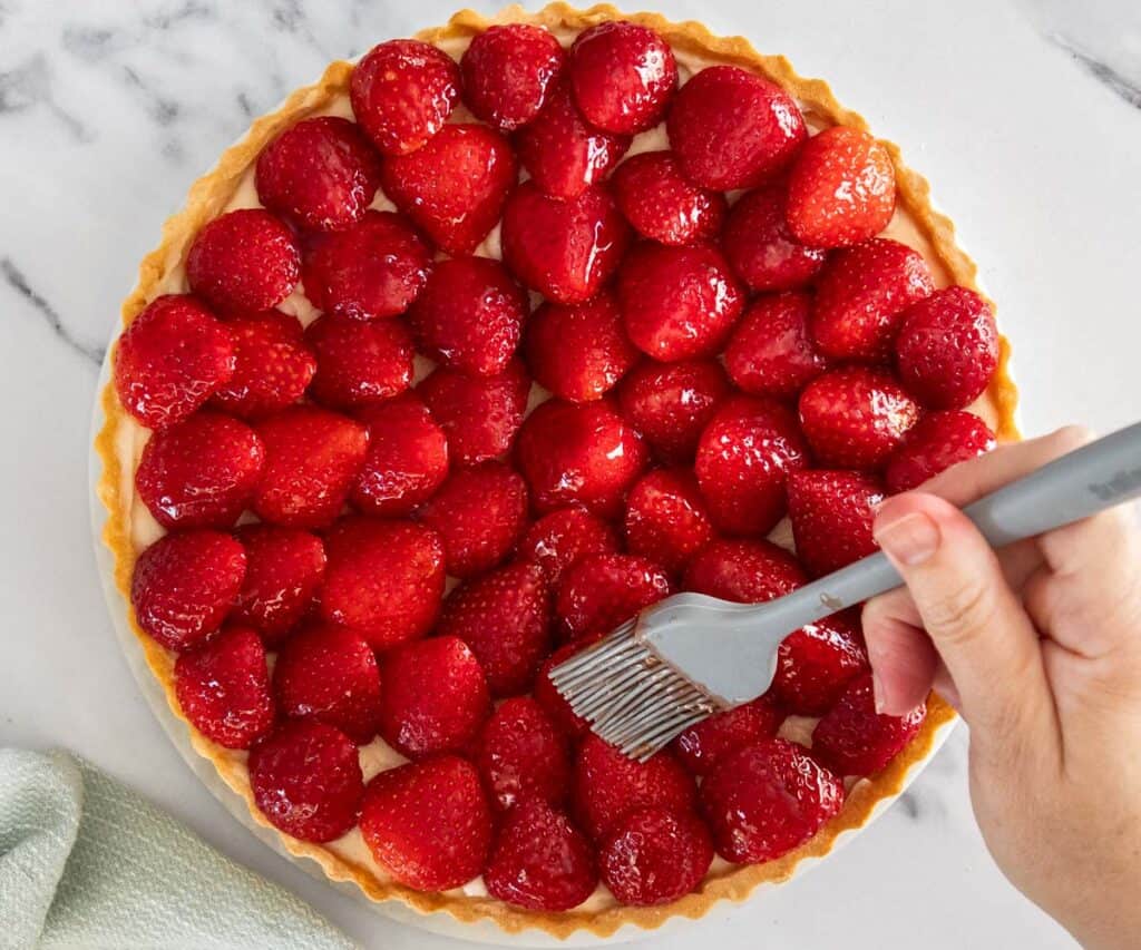 A hand using a pastry brush to brush jam over the strawberries on the tart.