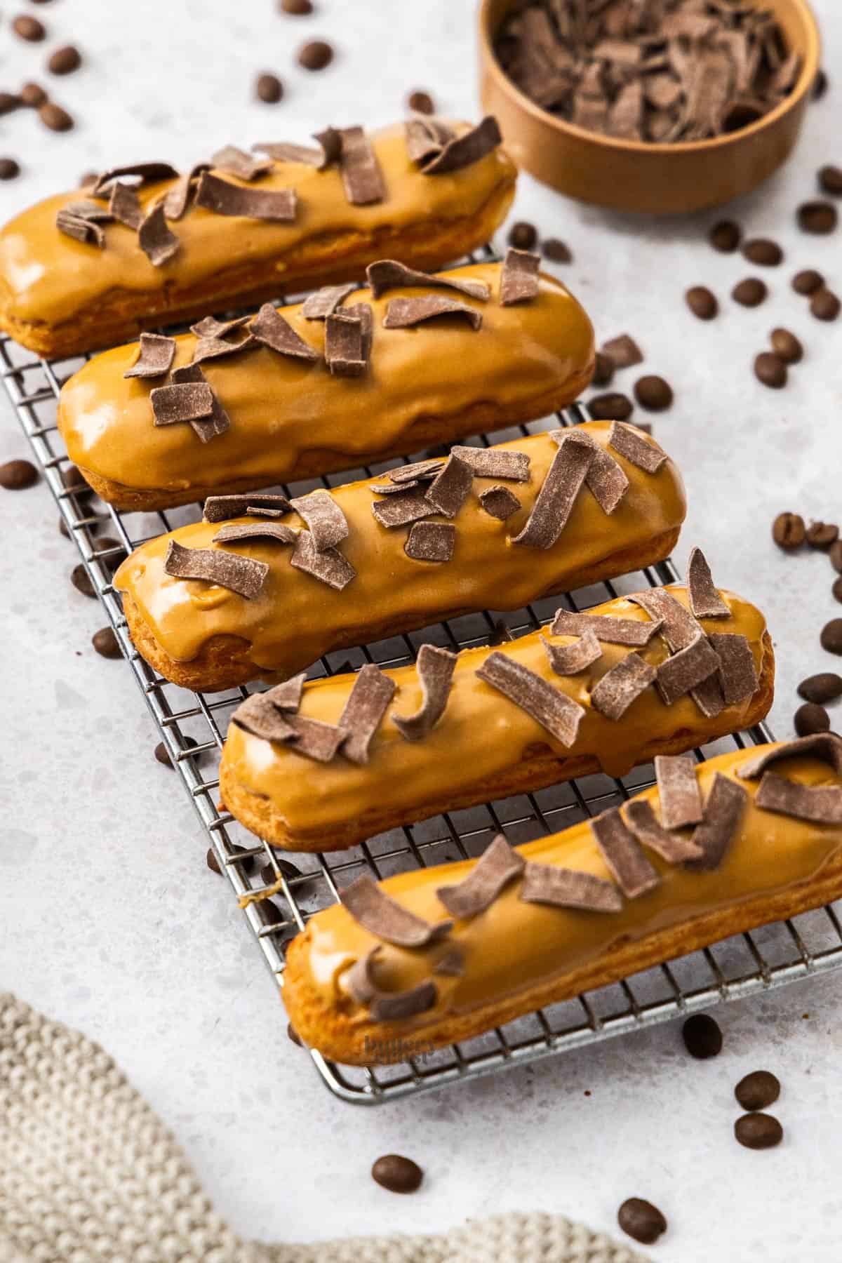 5 coffee eclairs topped with chocolate curls on a small wire rack.
