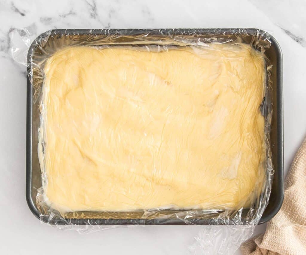 Pastry cream in a baking tray with a piece of plastic wrap pressed to the surface.