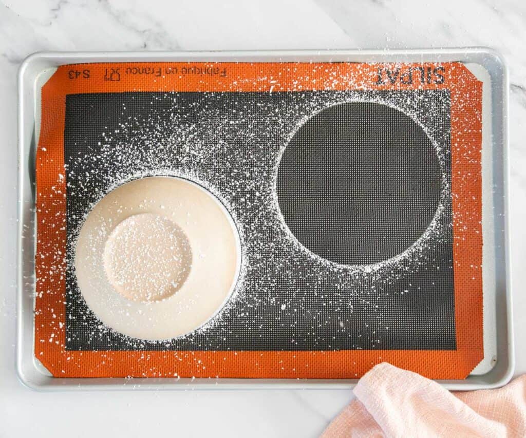 Top down view of a bowl upturned on a baking sheet with icing sugar dusted over it, a ring of sugar to the right where a bowl has been taken away.