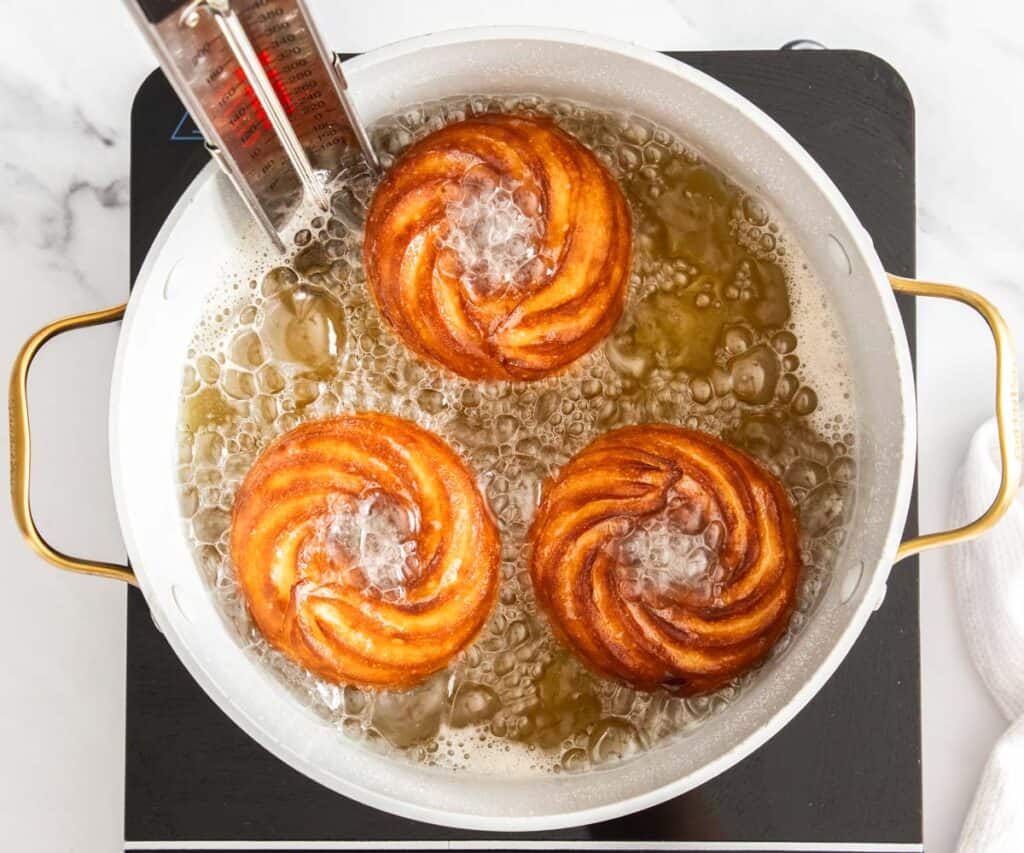 Three French cruller doughnuts looking golden in a pan of oil.