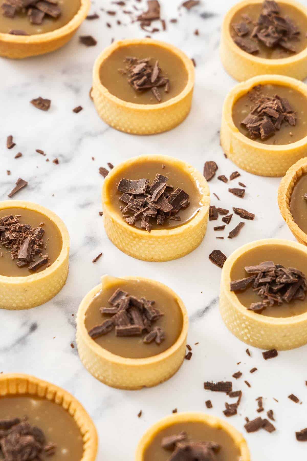 A batch of butterscotch tartlets topped with shaved chocolate on a marble surface.
