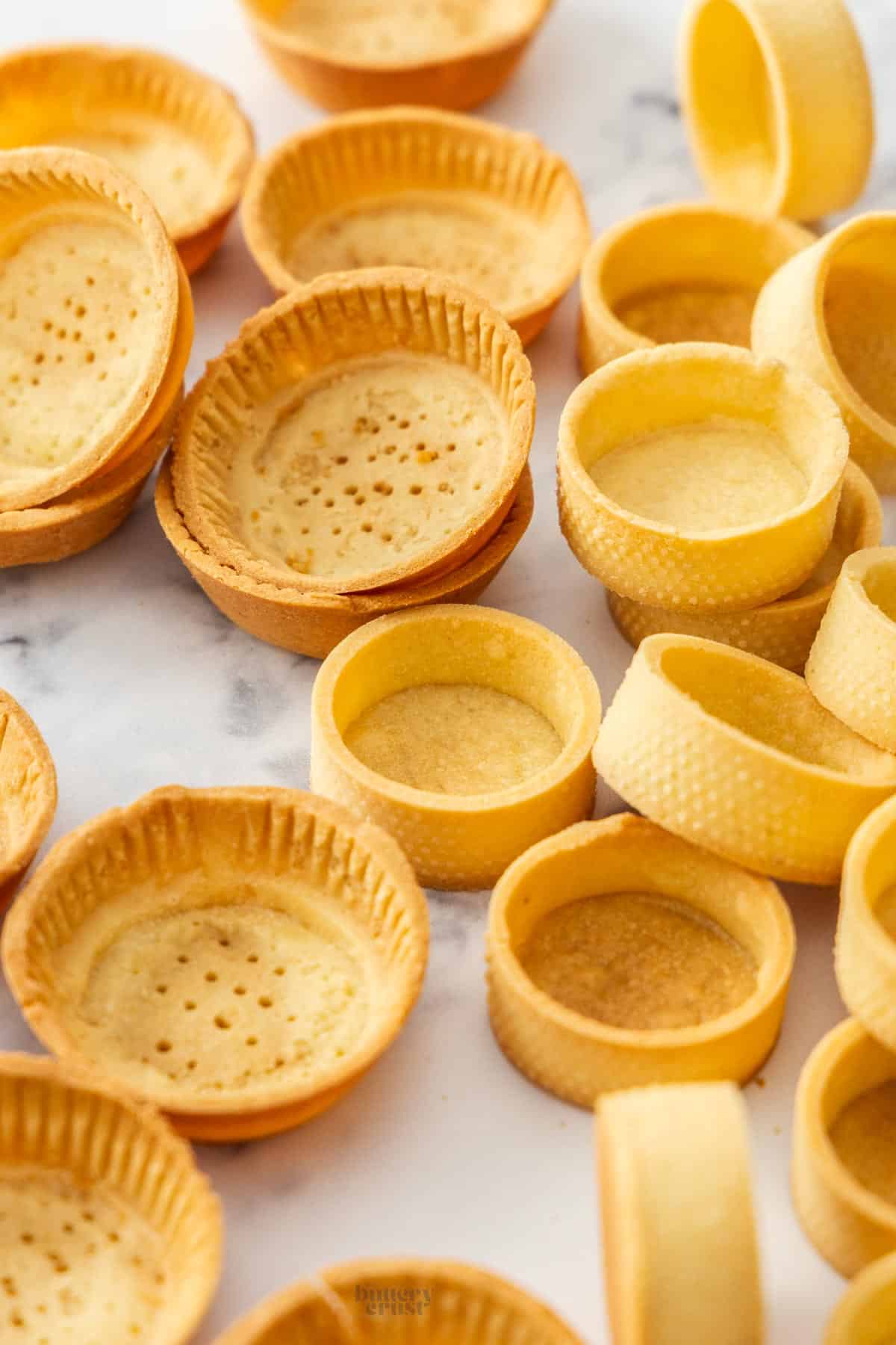 A pile of baked tartlet shells, some made in a muffin pan and some in tart rings.