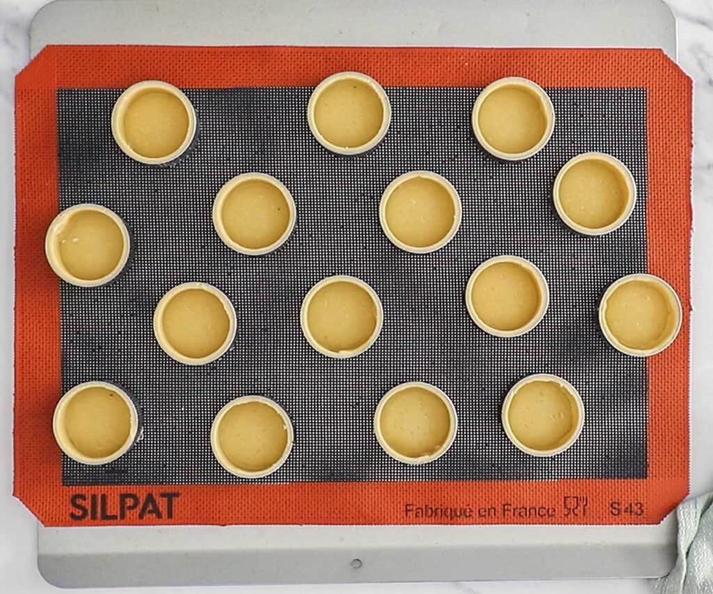 Fifteen mini tart shells in tart rings on a baking sheet.